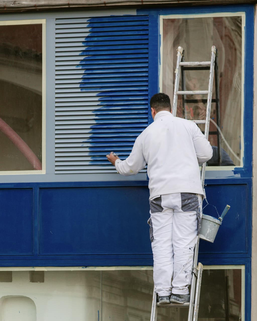 Painter in white work clothes on a ladder, halfway through painting a storefront shutter deep blue