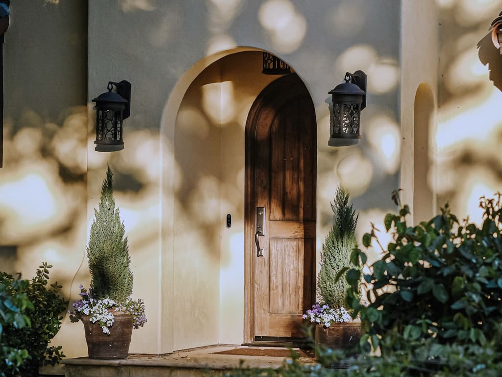 Arched stucco entry with a wooden front door, two wall lanterns and potted plants in soft late-afternoon light