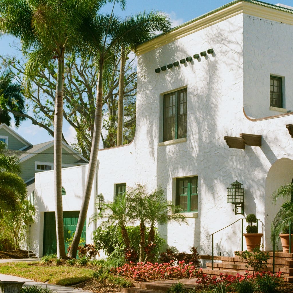 Two-story white stucco home with green-trimmed windows, wall lanterns, palm trees and tropical planting in bright daylight