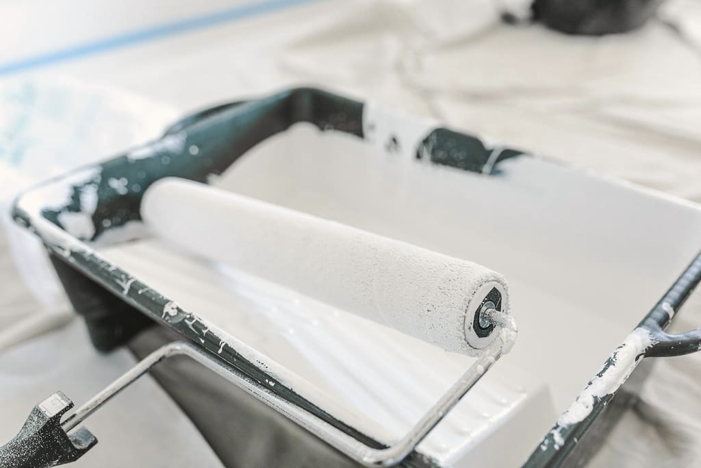 Paint roller loaded with white paint resting in a paint tray on a drop cloth