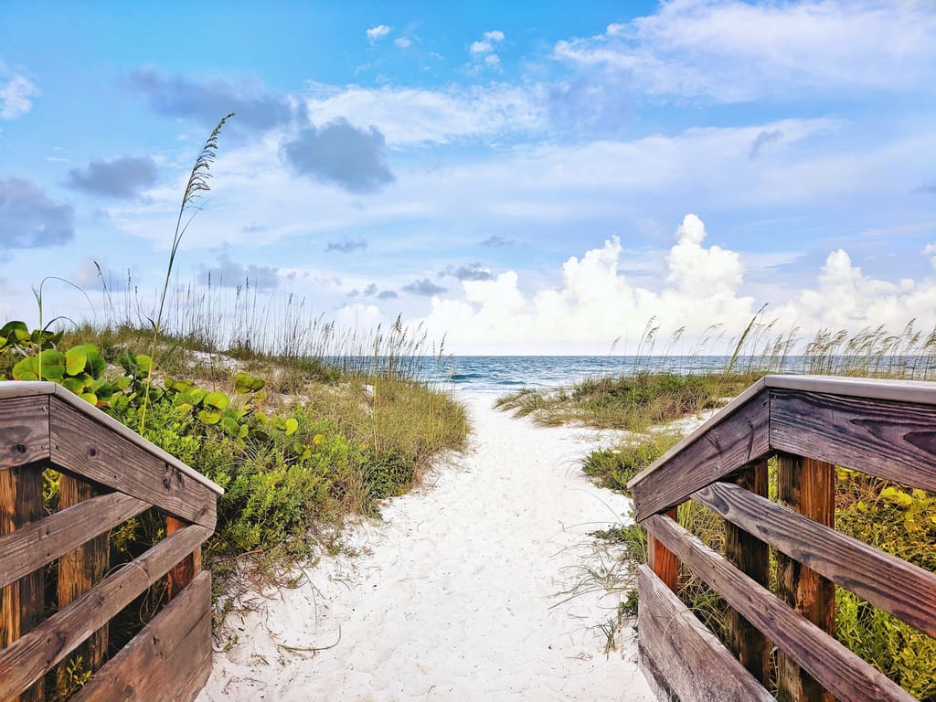 Weathered wooden beach walkway opening onto white sand, sea oats and calm blue water under a bright sky with scattered clouds