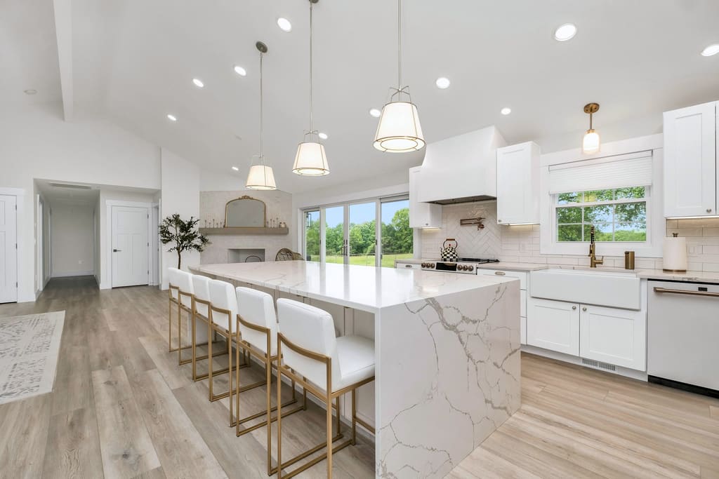 Bright kitchen with white shaker cabinets, a marble waterfall island, brass fixtures and light wood floors in daylight