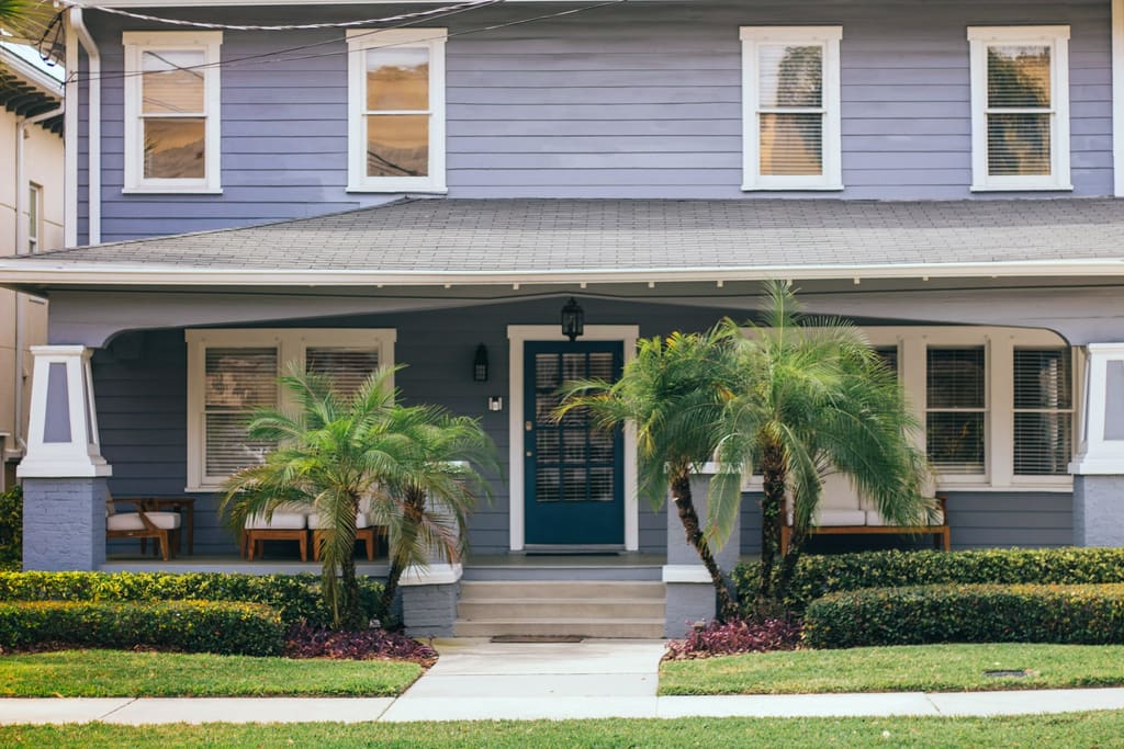 Two-story home painted soft blue-gray with crisp white trim, a navy front door, a deep front porch and palms along the walk