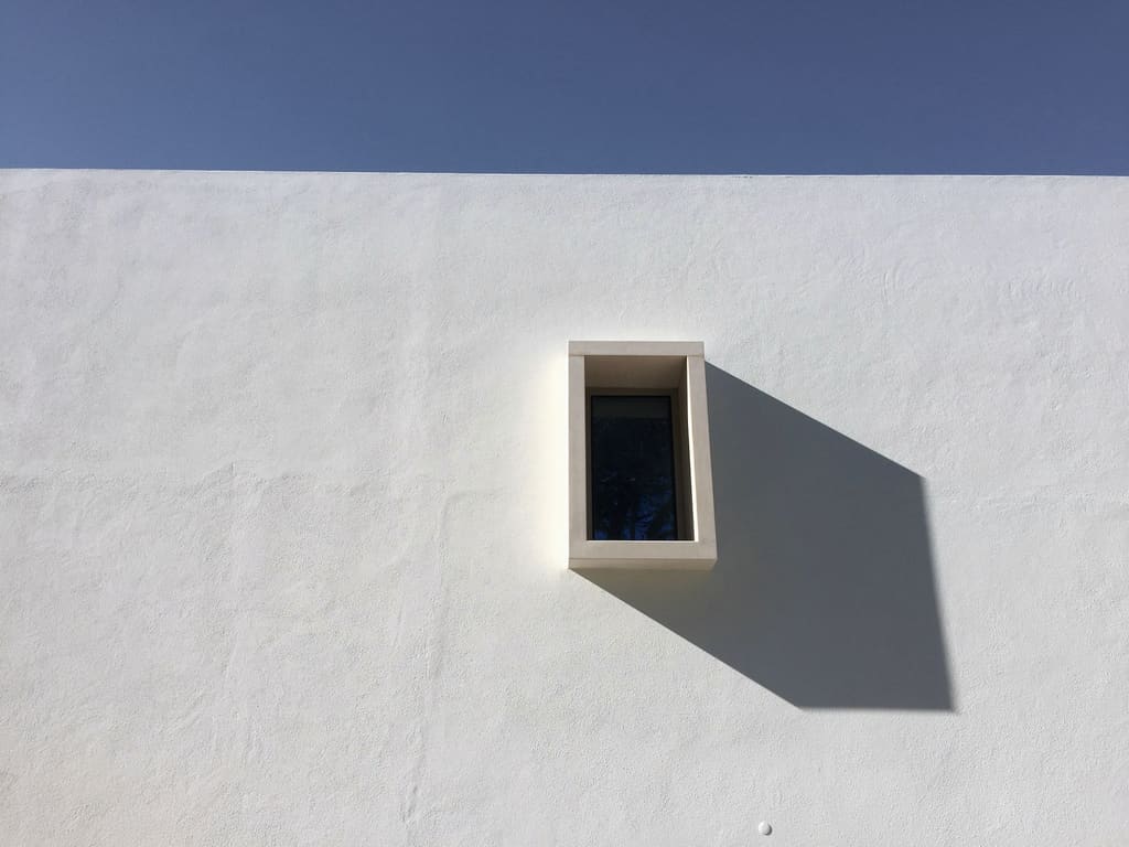 Smooth white stucco wall with a small recessed window casting a sharp shadow under a clear blue sky